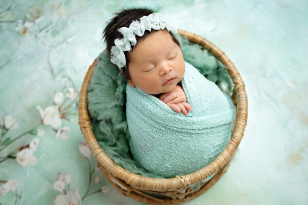 Baby girl wrapped in mint green inside wicker basket for studio newborn portraits