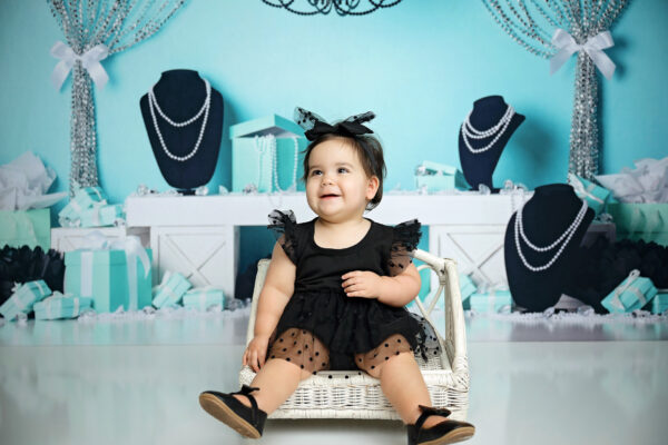 Smiling baby girl in a black dress and bow seated in a white wicker chair against a turquoise Breakfast at Tiffany's birthday backdrop.