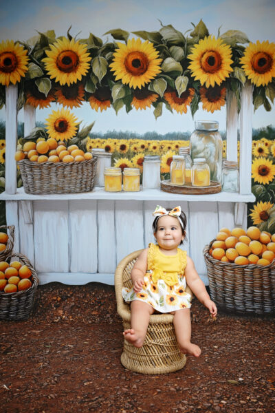 First birthday portrait of a baby girl in a yellow dress seated beneath a sunflower-filled market stand backdrop.