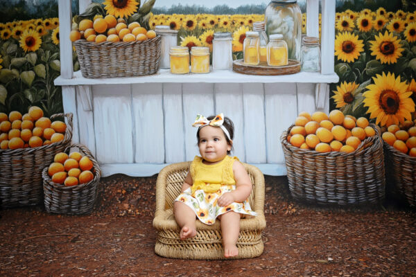 Baby girl wearing a yellow sunflower dress sitting in a wicker basket with a cheerful sunflower market backdrop.