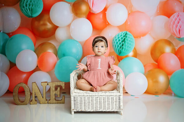 Baby girl sitting in a white chair with coral turquoise pink and gold balloons for her first birthday