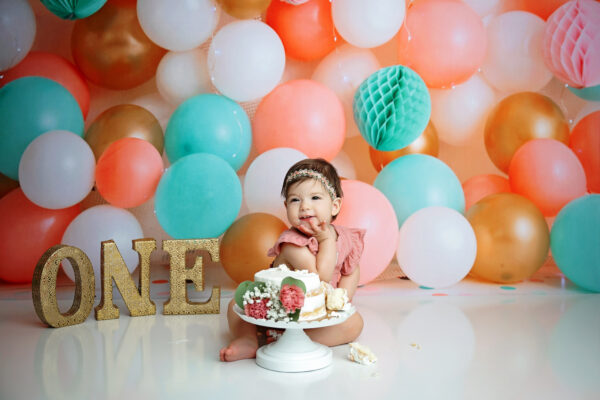 One-year-old girl posing beside her birthday cake during a colorful balloon cake smash in San Antonio