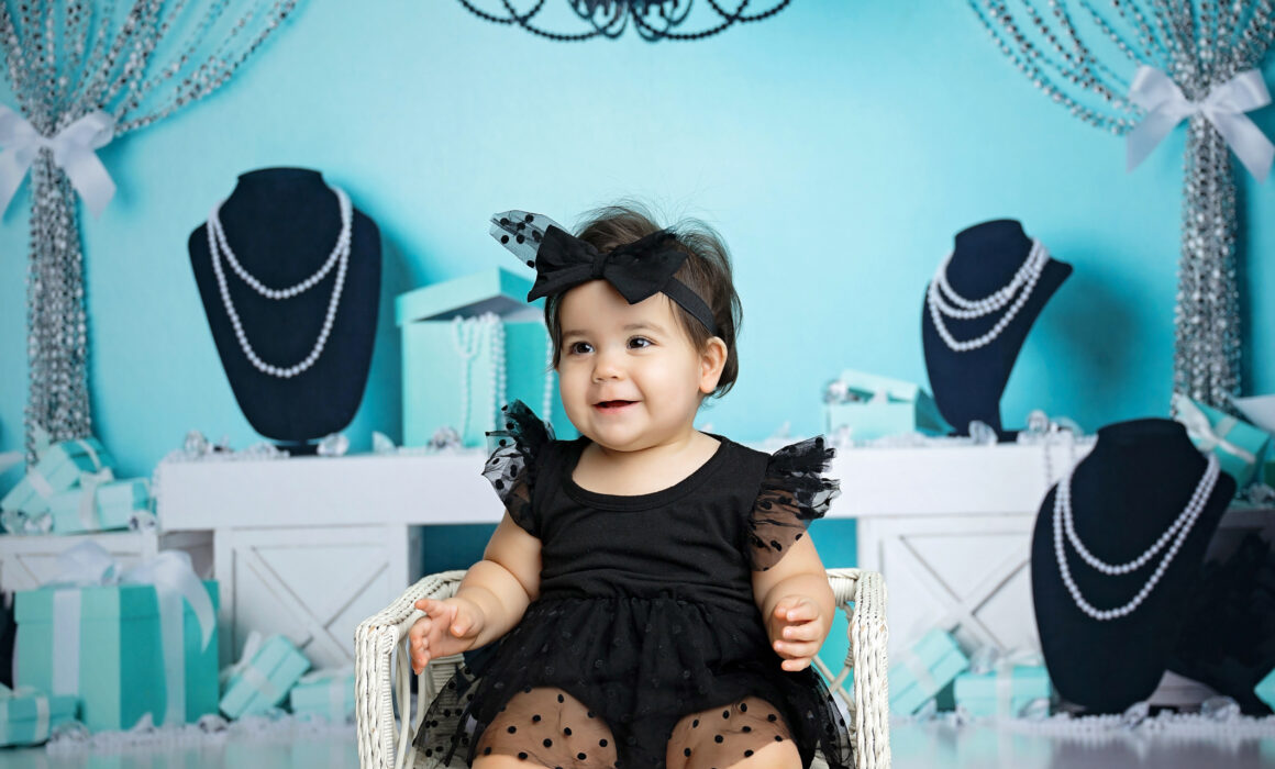 Close-up birthday portrait of a smiling baby girl in a black polka-dot tulle outfit with turquoise decorations behind her.