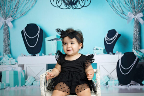 Close-up birthday portrait of a smiling baby girl in a black polka-dot tulle outfit with turquoise decorations behind her.