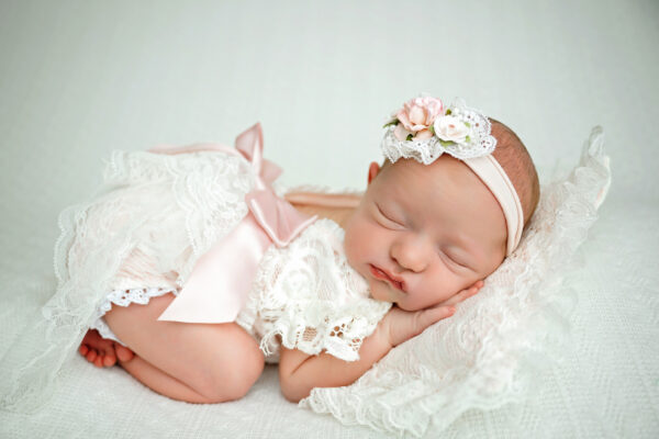 Sleeping newborn baby girl posed on a cream blanket wearing a delicate lace outfit and blush pink floral headband.