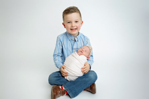 Smiling big brother sitting cross-legged and gently holding his swaddled newborn baby sister during a studio sibling photo session.