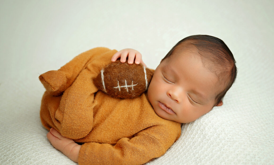 Sleeping newborn baby boy wearing a caramel knit outfit and holding a small felt football on a cream blanket.