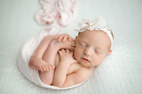 Peacefully sleeping newborn girl wrapped in soft blush pink fabric with a white lace bow headband during her newborn photo session.