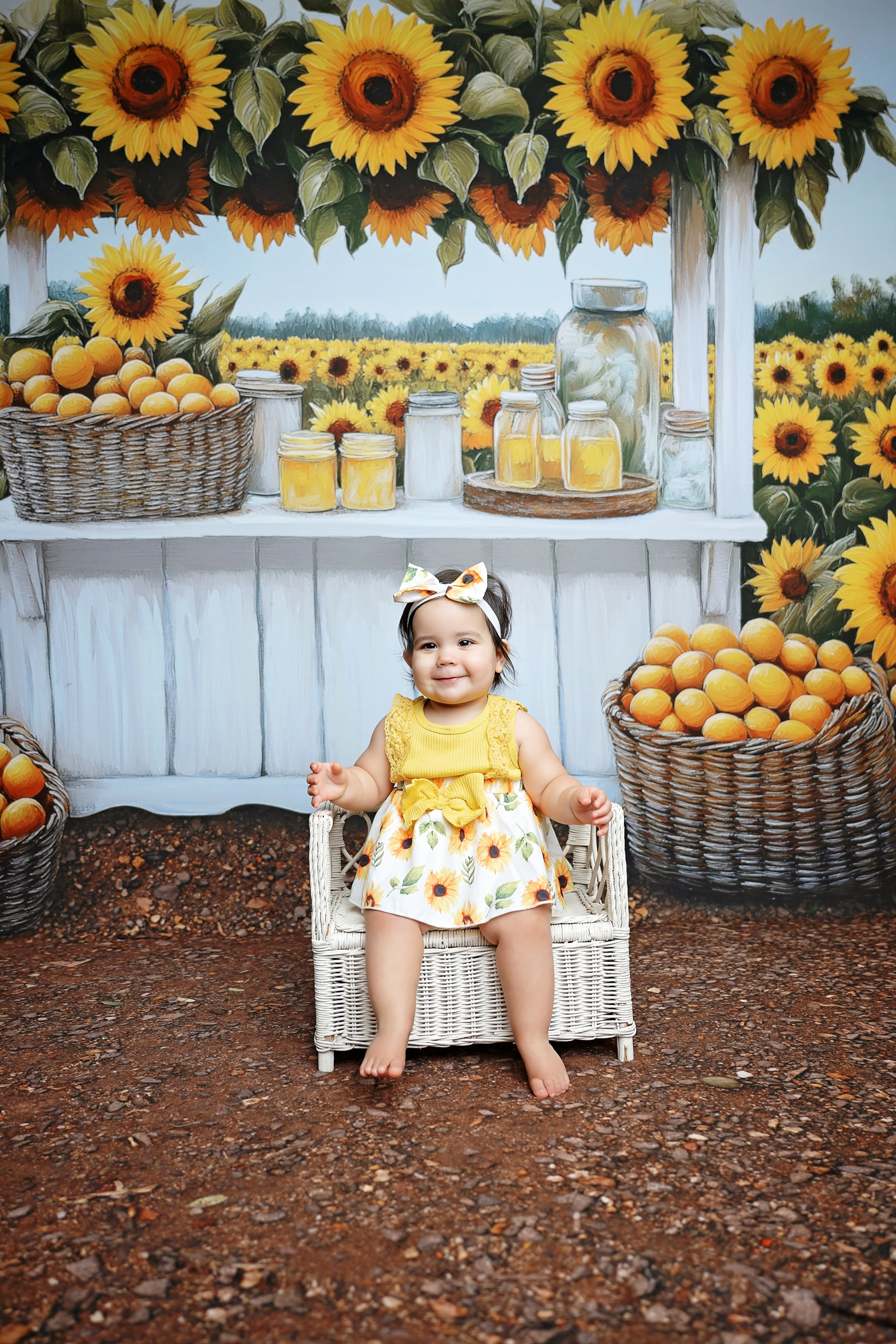 Baby girl looking upward with a sweet smile while seated in a wicker chair against a sunflower market backdrop.