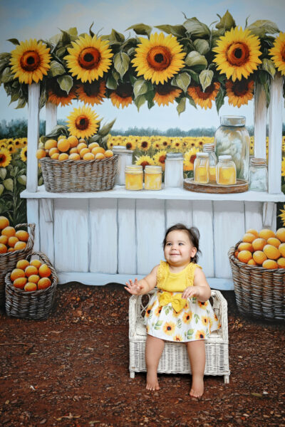Smiling baby girl in a yellow sunflower dress sitting in a white wicker chair during her first birthday milestone session.