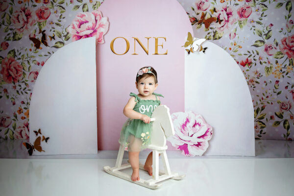 One-year-old girl standing with a white rocking horse during a floral milestone photography session