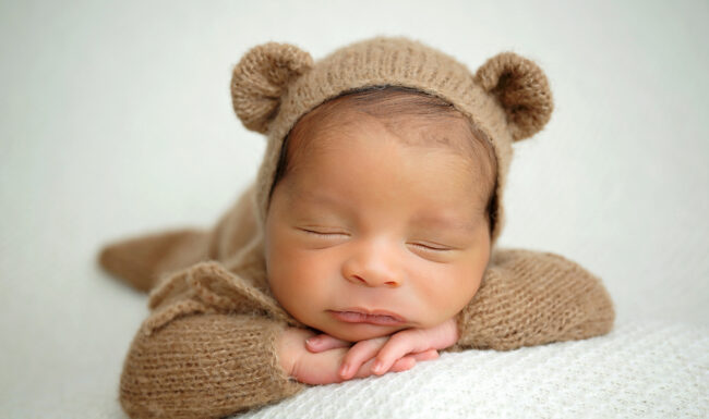 Close-up newborn portrait of a sleeping baby wearing a soft brown teddy bear bonnet with hands resting under his chin.